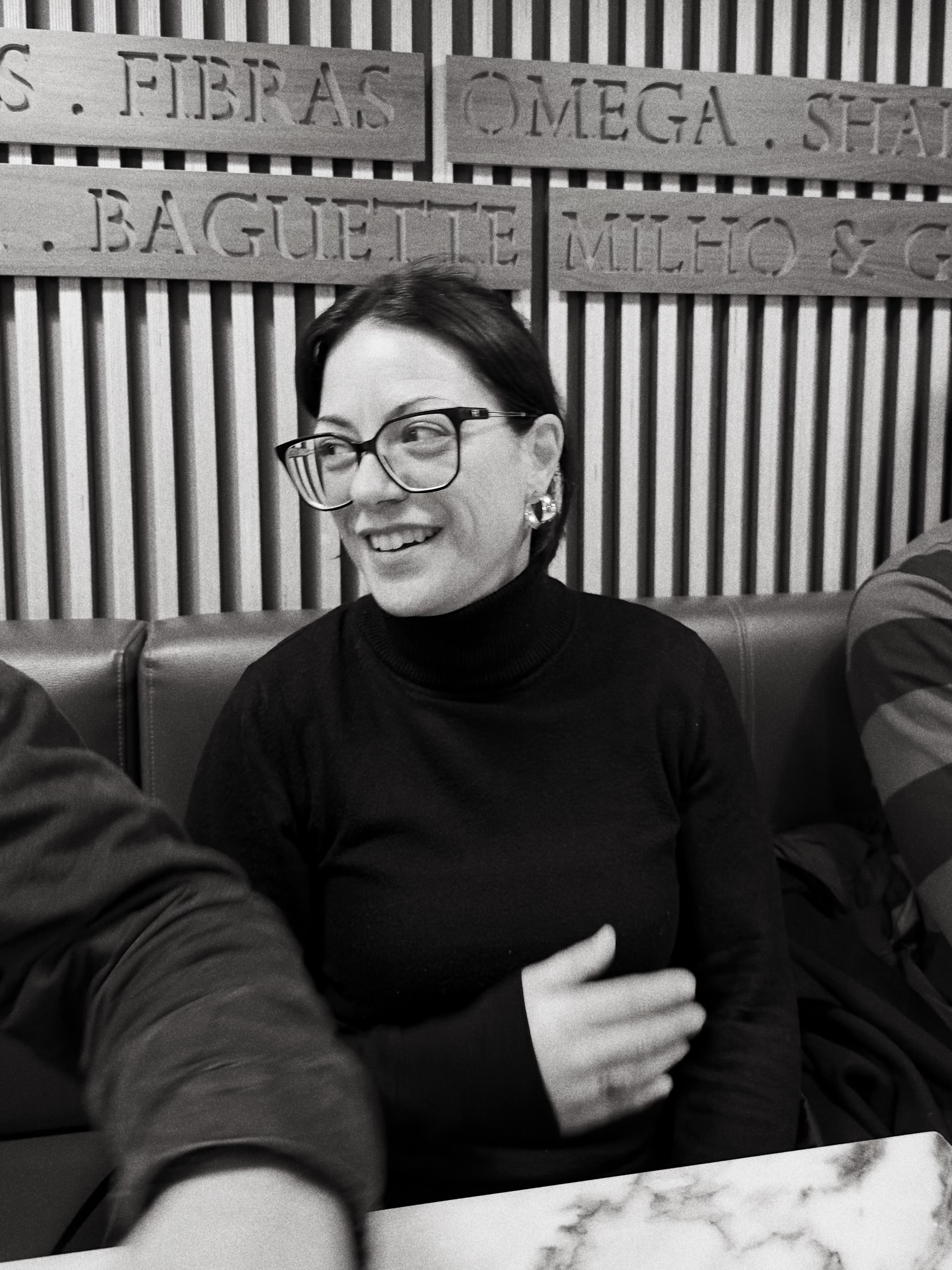 Uma mulher com óculos e gola alta preta, a sorrir, sentada numa mesa de café.