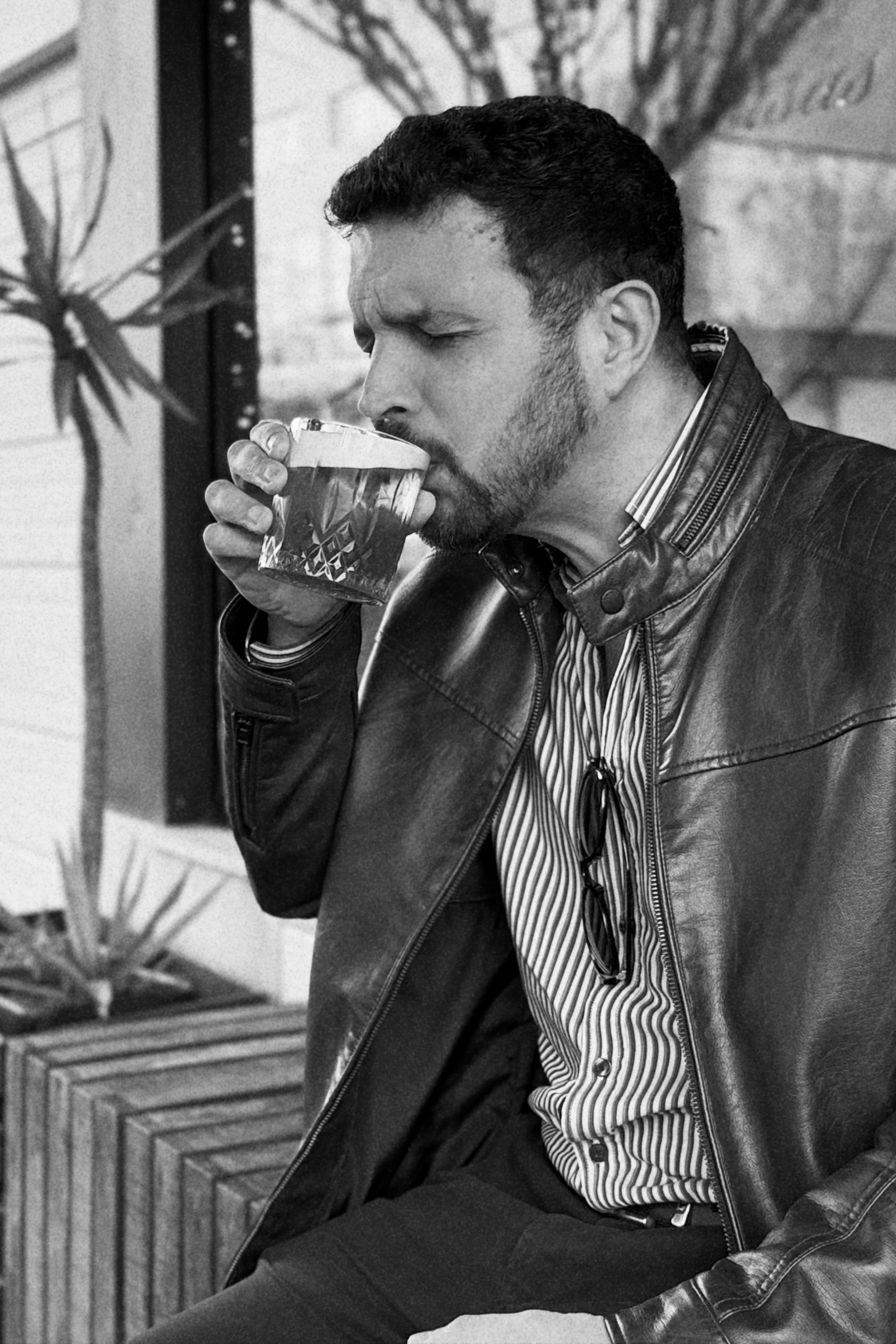 Homem barbudo a beber de um copo, vestido com casaco de cabedal e camisa às riscas, em preto e branco.