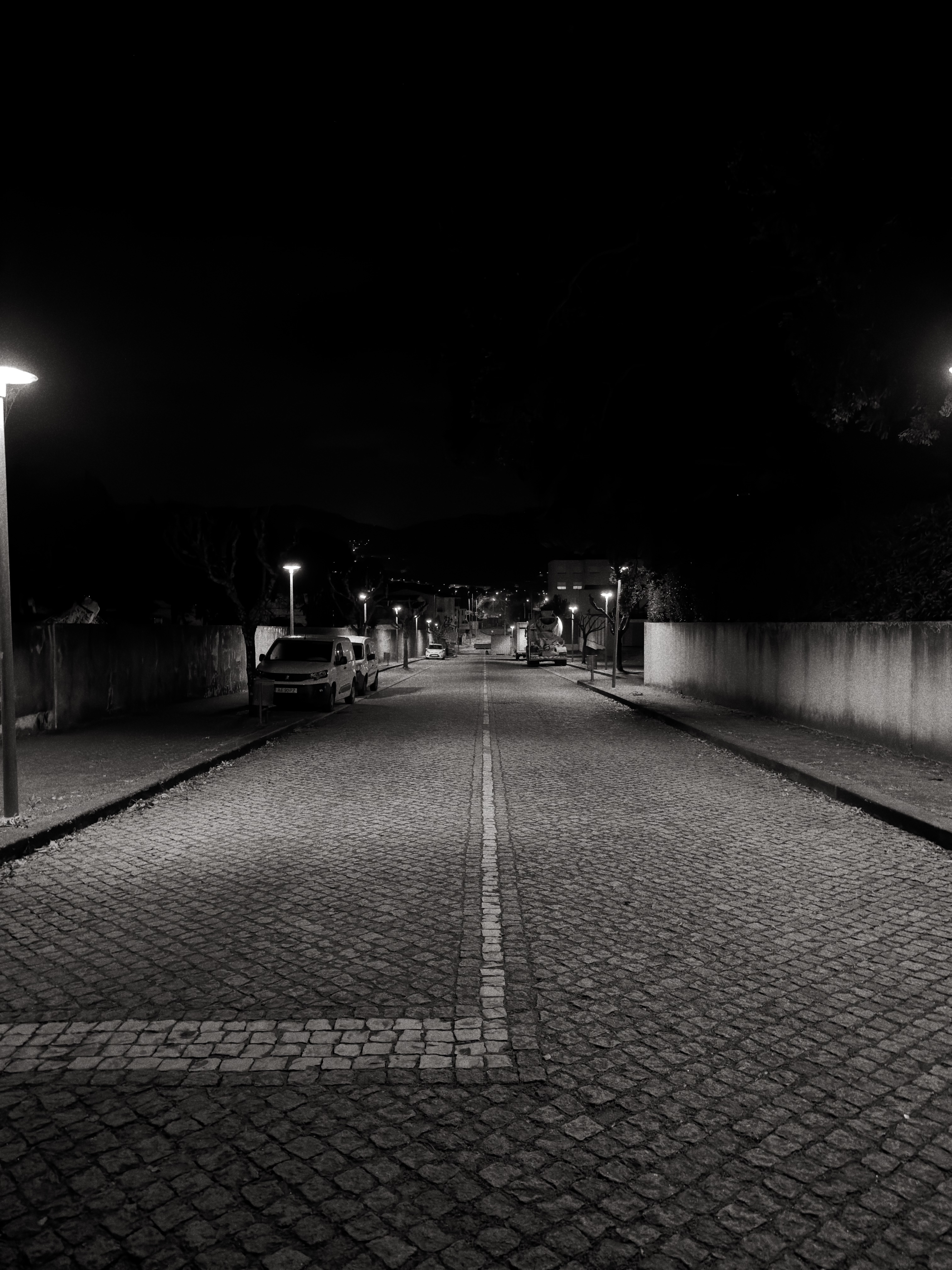 Fotografia a preto e branco de uma rua de calçada vazia à noite, iluminada por candeeiros.